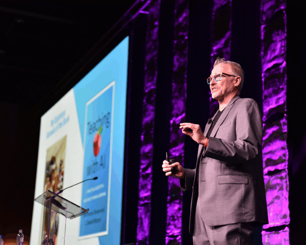 A photo of the keynote speaker from OLC Accelerate 2024 giving his presentation from an elevated stage. He stands on an elevated stage, in front of a large projection of a slide show.